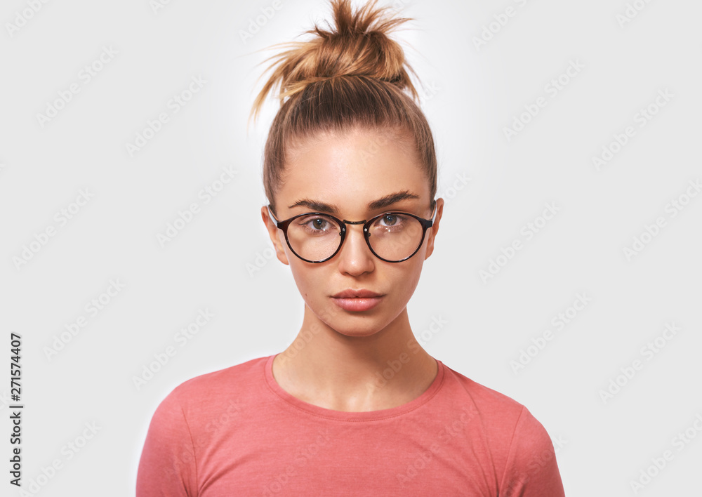 Portrait of attractive blonde young woman wearing round transparent spectacles and casual pink blouse, looks seriously directly into camera, poses against white studio background. People and emotions