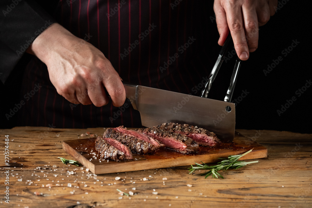 Slicing juicy beef steak by knife in chef hands closeup. Food cooking concept. Dark black background copy space.