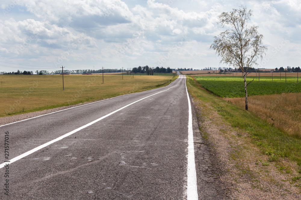View of the small asphalt road in the countryside with blue sky and cumulus clouds on the background.