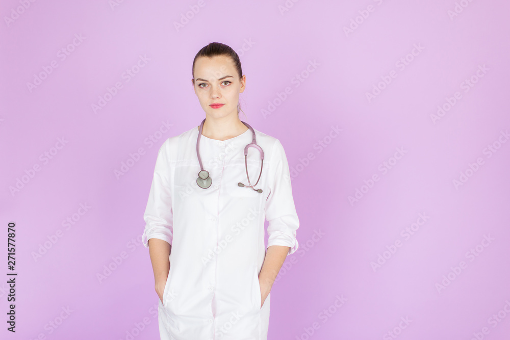 Young female blonde doctor with her hands in pockets with stethoscope on her neck on pink background with space for text. Doctor looks at camera Young female blonde doctor with her hands in pockets with stethoscope on her neck on pink background with space for text. Doctor looks at camera