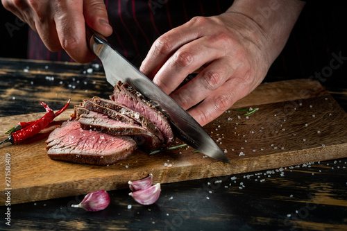Slicing juicy beef steak by knife in chef hands closeup. Food cooking concept. Dark black background copy space. photo