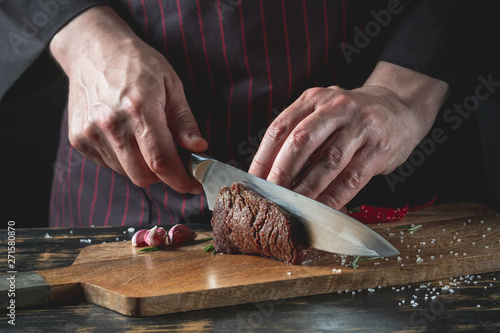 Slicing juicy beef steak by knife in chef hands closeup. Food cooking concept. Dark black background copy space.