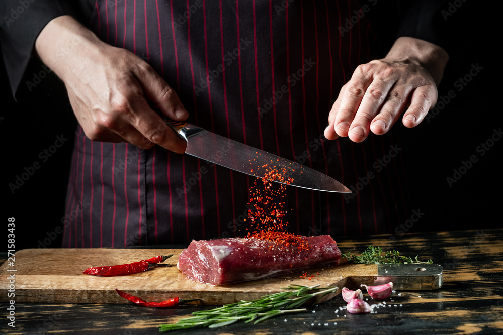 Butcher chef cooking meat fillet and adding salt, pepper and chili for marinade on black background.