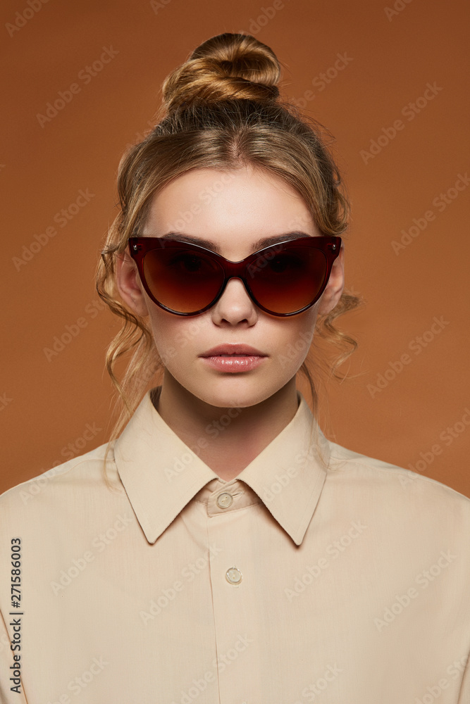 Cropped front view shot of blonde lady, wearing shirt. The girl with bun and wavy hair locks in butterfly-shaped sunglasses with brown rim and lenses, is looking at camera against brown background. Cropped front view shot of blonde lady, wearing shirt. The girl with bun and wavy hair locks in butterfly-shaped sunglasses with brown rim and lenses, is looking at camera against brown background.