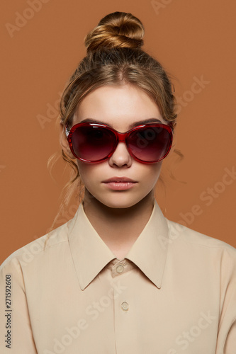 Cropped front view shot of lady with fair hair, wearing beige shirt. The girl with bun and wavy hair locks in oval-shaped sunglasses with red rim, is looking at camera on the light brown background.