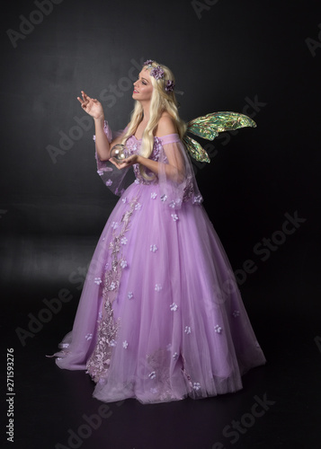 full length portrait of a blonde girl wearing a fantasy fairy inspired costume, long purple ball gown with fairy wings, standing pose holding a crystal ball on a dark studio background.