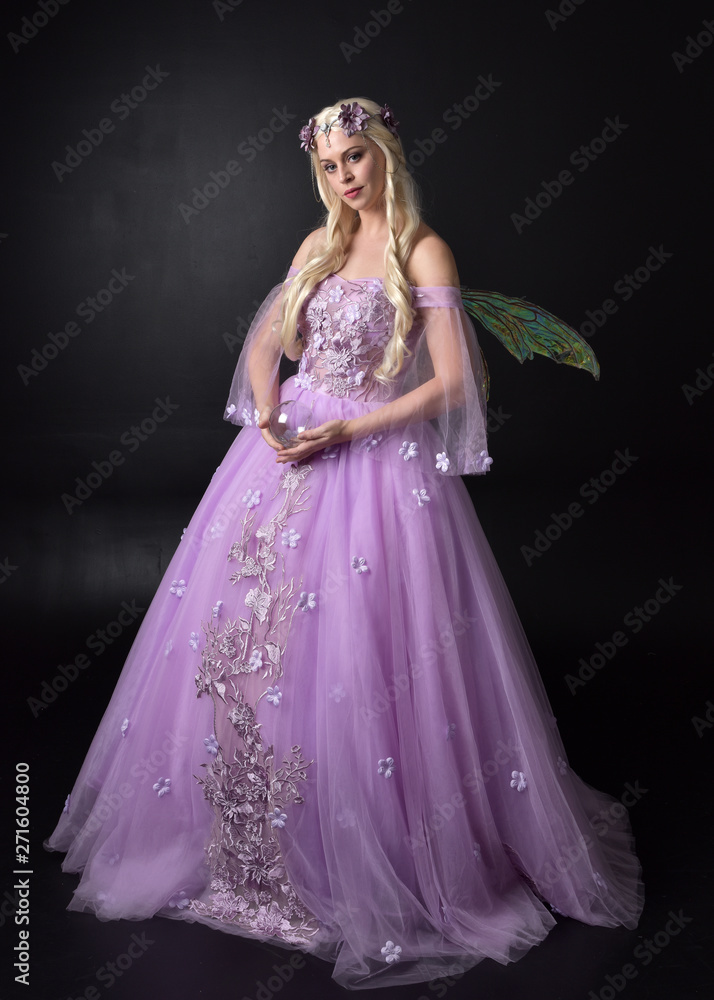 full length portrait of a blonde girl wearing a fantasy fairy inspired costume,  long purple ball gown with fairy wings,  standing pose holding a crystal ball on a dark studio background.