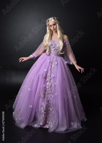 full length portrait of a blonde girl wearing a fantasy fairy inspired costume   long purple ball gown with fairy wings    standing pose on a dark studio background.