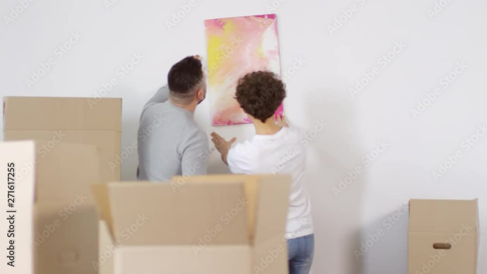 Panning rear shot of smiling Caucasian couple putting up abstract painting onto white wall together, then trying to straighten it, with plenty of cardboard boxes all around their new home Panning rear shot of smiling Caucasian couple putting up abstract painting onto white wall together, then trying to straighten it, with plenty of cardboard boxes all around their new home