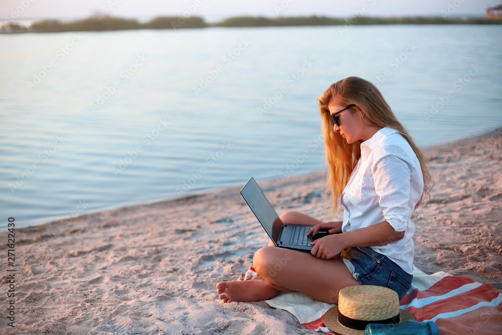 Authentic woman working with laptop on sea shore. Freelancer girl telecommuting with team on project on tropical island. Female using internet from computer while sitting on beach towel on sunset.