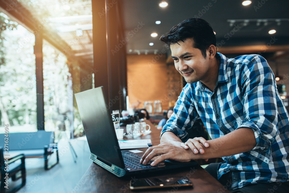 Attractive asian male is typing messages on a laptop while sitting against the wide windows of a coffee shop. a hipster girl is working on a portable computer in a cafe, freelance man. Attractive asian male is typing messages on a laptop while sitting against the wide windows of a coffee shop. a hipster girl is working on a portable computer in a cafe, freelance man.