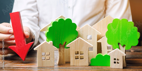 Businessman holds down arrow near wooden houses and trees. The concept of reducing investment in the construction of new buildings. Drop in demand for home purchase. Illegal construction of park areas