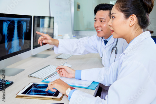 Asian male doctor pointing at computer monitor with x-ray image and discussing some problems with his colleague they working at the office photo