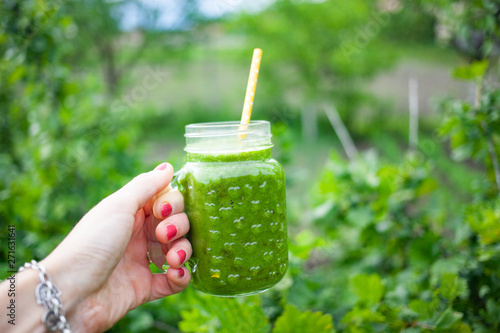 woman's hand holding jar with green cold-pressed juice, vegetable garden background. Healthy eating, detoxing, juicing, body cleancing concept photo