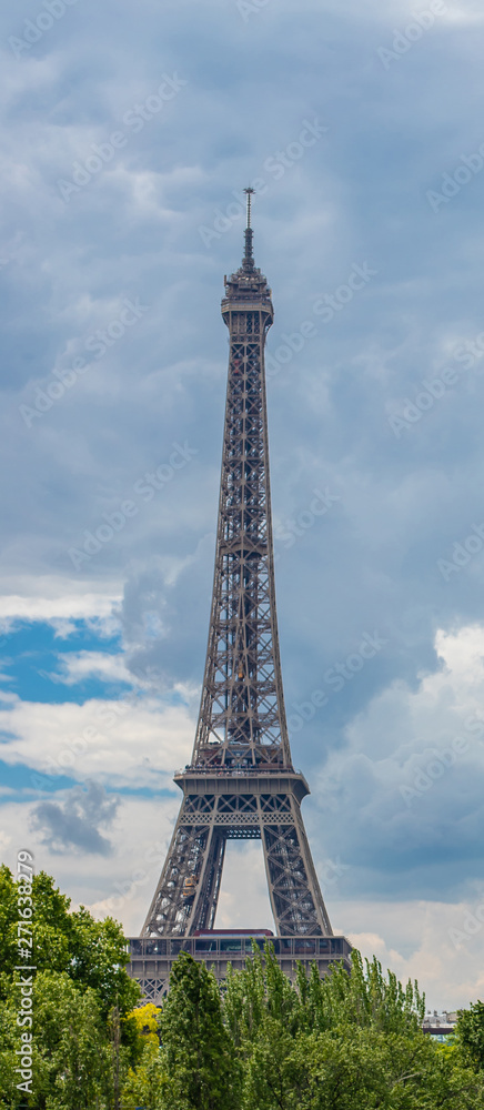 Fototapeta premium Eiffel Tower behind the trees against the backdrop of a bright cloudy sky.