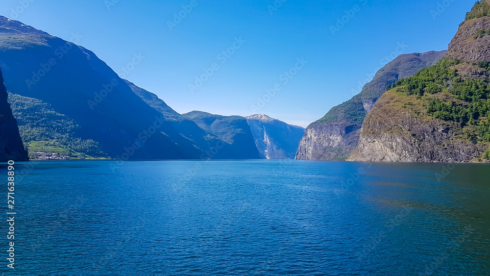 A view on the Songefjorden (King of the Fjords) from the water level. It is the deepest fjord in Norway. Tall, lush green mountains surrounding the fjord. Calm surface of the water. Clear blue sky. A view on the Songefjorden (King of the Fjords) from the water level. It is the deepest fjord in Norway. Tall, lush green mountains surrounding the fjord. Calm surface of the water. Clear blue sky.