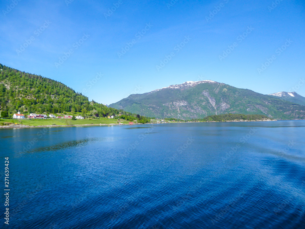 A view on the Songefjorden (King of the Fjords) from the water level. It is the deepest fjord in Norway. Tall, lush green mountains surrounding the fjord. Wavy surface of the water. Clear blue sky.