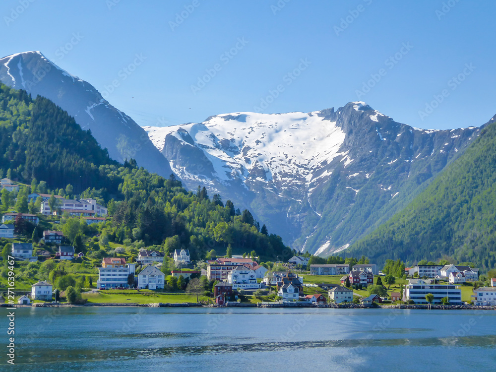 A little village located at the bottom of a fjord, just on the coast line. Tall mountains surrounding the village are covered with snow. Clear and bright day. Calm surface of the water.