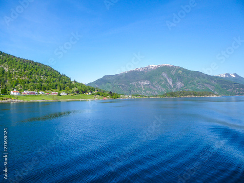 A view on the Songefjorden (King of the Fjords) from the water level. It is the deepest fjord in Norway. Tall, lush green mountains surrounding the fjord. Wavy surface of the water. Clear blue sky.