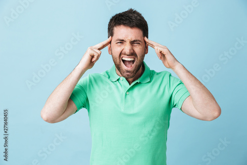 Stressed young handsome bearded man posing isolated over blue wall background screaming because of headache.