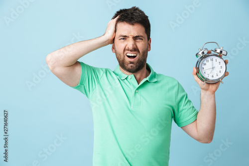 Shocked confused young handsome bearded man posing isolated over blue wall background holding alarm clock.