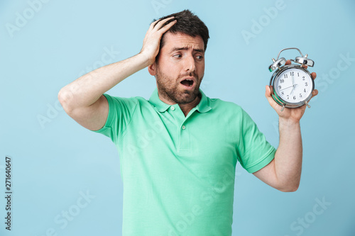 Shocked confused young handsome bearded man posing isolated over blue wall background holding alarm clock.