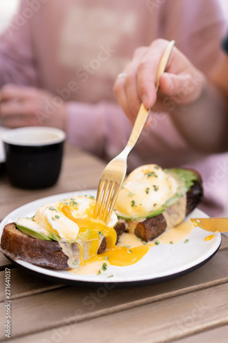 Morning breakfast with friends in a cozy summer outdoor cafe. Poached eggs on toasted black bread with avocado and Dutch sauce. Hands in the frame with golden cutlery.