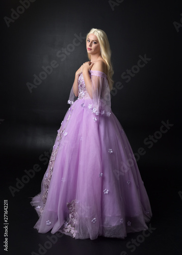 full length portrait of a blonde girl wearing a fantasy fairy inspired costume   long purple ball gown with fairy wings    standing pose  on a dark studio background.