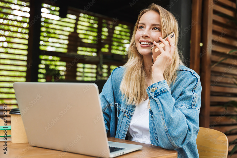 Excited happy young woman posing outdoors in cafe using laptop computer and talking by mobile phone.