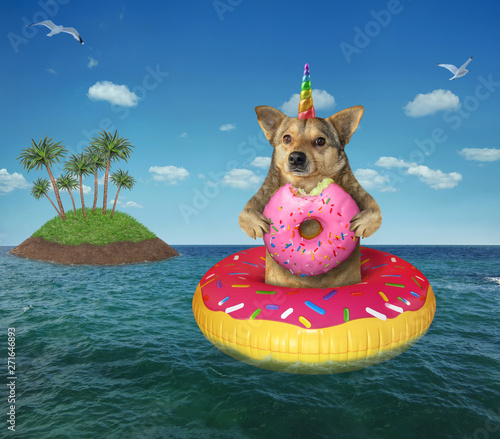 The dog unicorn with a pink donut on an inflatable circle drifts past an island on the high seas. © iridi66