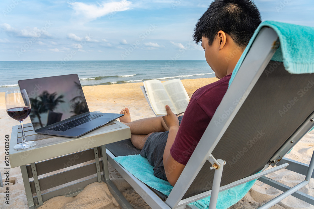 Asian man reading book next to laptop on beautiful beach Asian man reading book next to laptop on beautiful beach