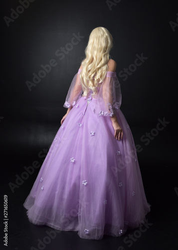 full length portrait of a blonde girl wearing a fantasy fairy inspired costume, long purple ball gown with fairy wings, standing pose on a dark studio background.