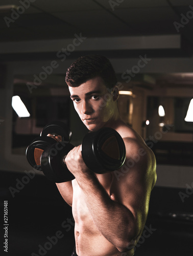 Strong young guy covered with electric discharges looking at camera while lifting heavy dumbbells in gym
