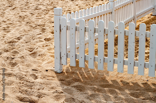 Retro style white painted wooden fence on beach sand with clear striped shadows. Minimal summer seascape. Uneven surface of clear sand. Summer sunny day. Coastal landscape with white striped fence 