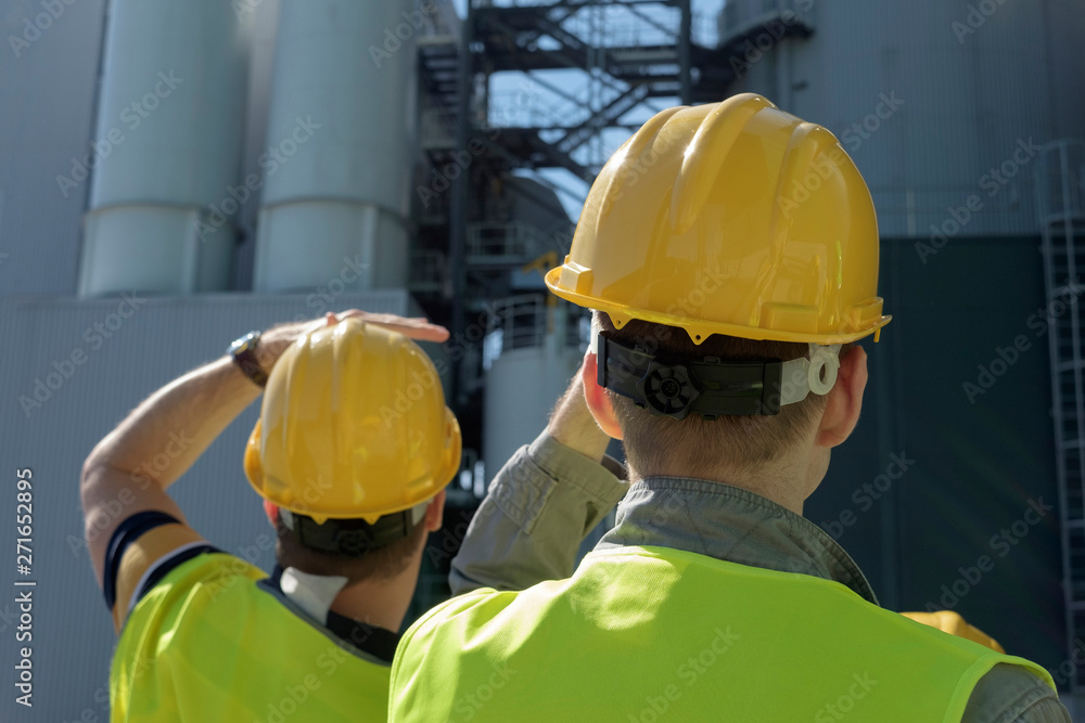 Two workers in yellow helmets and vests are discussing looking at an industrial object. Two workers in yellow helmets and vests are discussing looking at an industrial object.