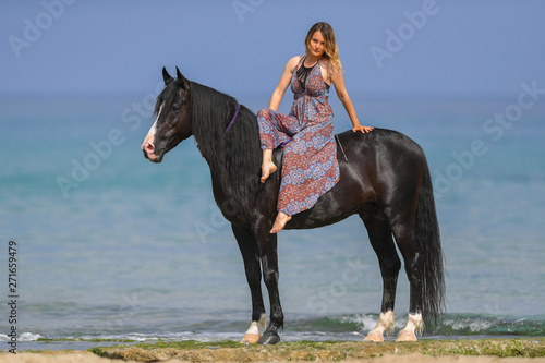Portrait of a beautiful blond girl sitting on the back of her black horse on the sea beach
