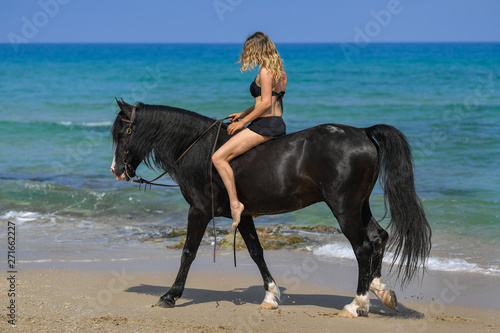 Portrait of a beautiful blond girl sitting on the back of her black horse on the sea beach