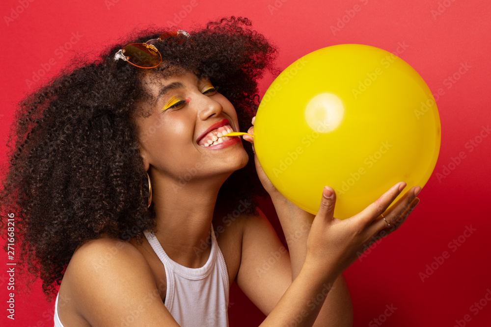 Mulatto model with a lush hairstyle of curly black hair inflates a yellow  balloon on a red Studio background Stock Photo | Adobe Stock