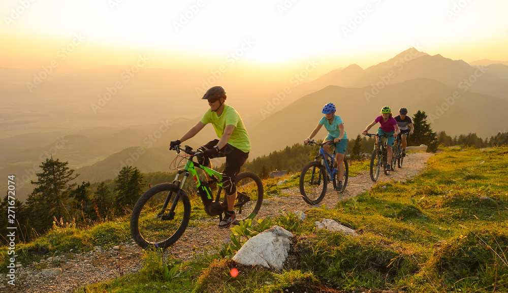 SUN FLARE Cheerful tourists ride electric bicycles up a mountain trail at sunset SUN FLARE Cheerful tourists ride electric bicycles up a mountain trail at sunset