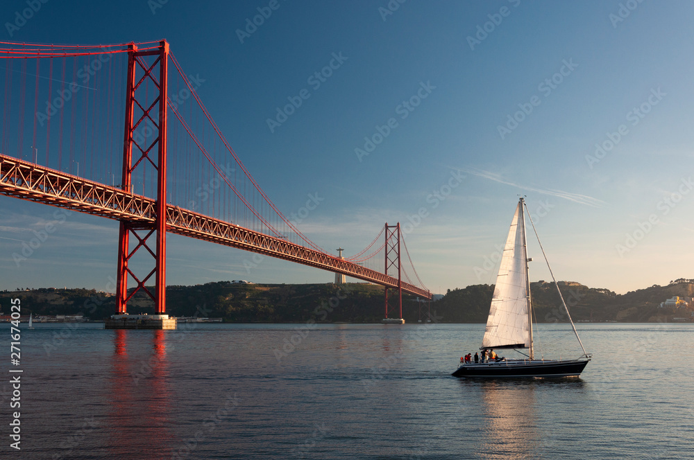 Sailing boat passing by the 25 of April Bridge (Ponte 25 de Abril) over the Tagus River in the city of Lisbon, Portugal; Concept for travel in Portugal