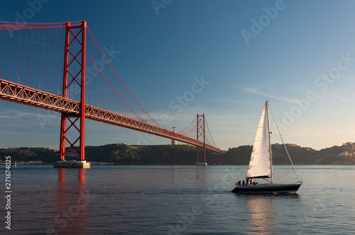 Sailing boat passing by the 25 of April Bridge (Ponte 25 de Abril) over the Tagus River in the city of Lisbon, Portugal; Concept for travel in Portugal