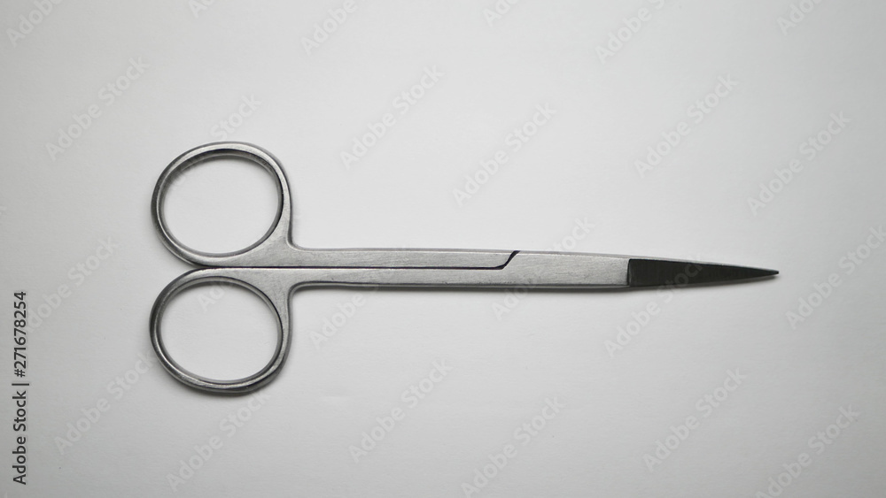 A pair of small stainless steel surgical medical grade scissors laying closed on a white table. A pair of small stainless steel surgical medical grade scissors laying closed on a white table.