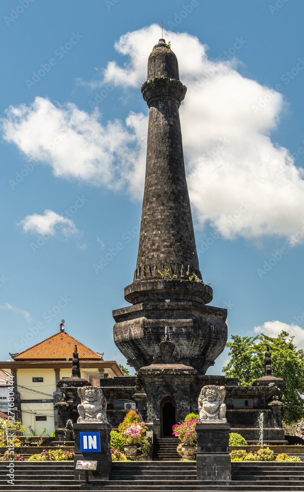 Klungkung, Bali, Indonesia - February 26, 2019: Black Oblisk Puputan Monument remembering a suicidal royal battle against invaders under blue sky. Phallus symbol and two lion statues as guards. Pink f