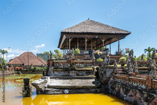 Klungkung, Bali, Indonesia - February 26, 2019: Brown stone Floating Pavilion at Royal Palace of Klungkung Kingdom with gray reed roof under blue sky. Mold covered monuments and yellow-brown lotus pon photo