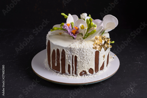 Chocolate cake decorated with flowers and poured white icing on black stone background.