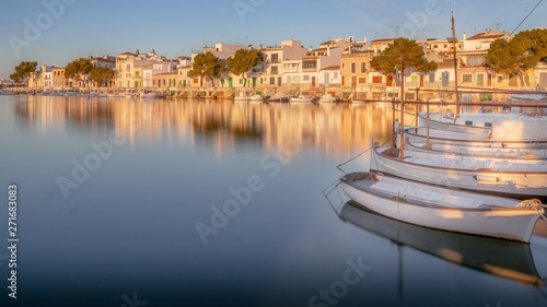Portocolom town houses, harbour, port, golden light, sunrise, calm blue mediterranean sea, fishing boats, sandy beach, reflection, blue sky, Mallorca, Spain. © gaz