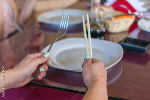 Female hand is holding chopsticks and fork over an empty plate.