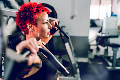 Young beautiful woman with short red hair at the gym by the shoulder press machine resting at workout bodybuilding fitness training