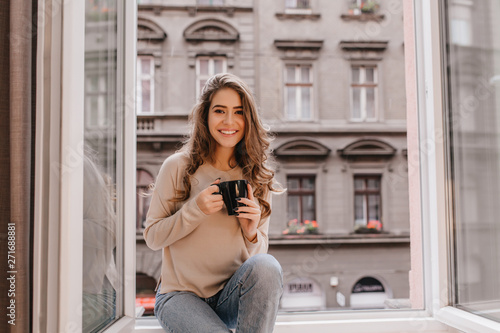 Картината върху платно Adorable woman expressing positive emotions while posing on sill with cup of latte