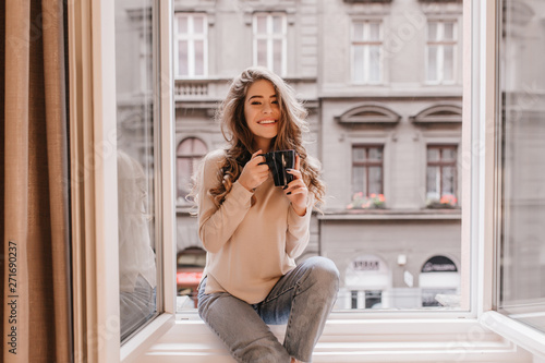 Happy white woman with gorgeous hairstyle sitting on window sill. Indoor portrait of glad young lady in jeans posing emotionally while drinking coffee.